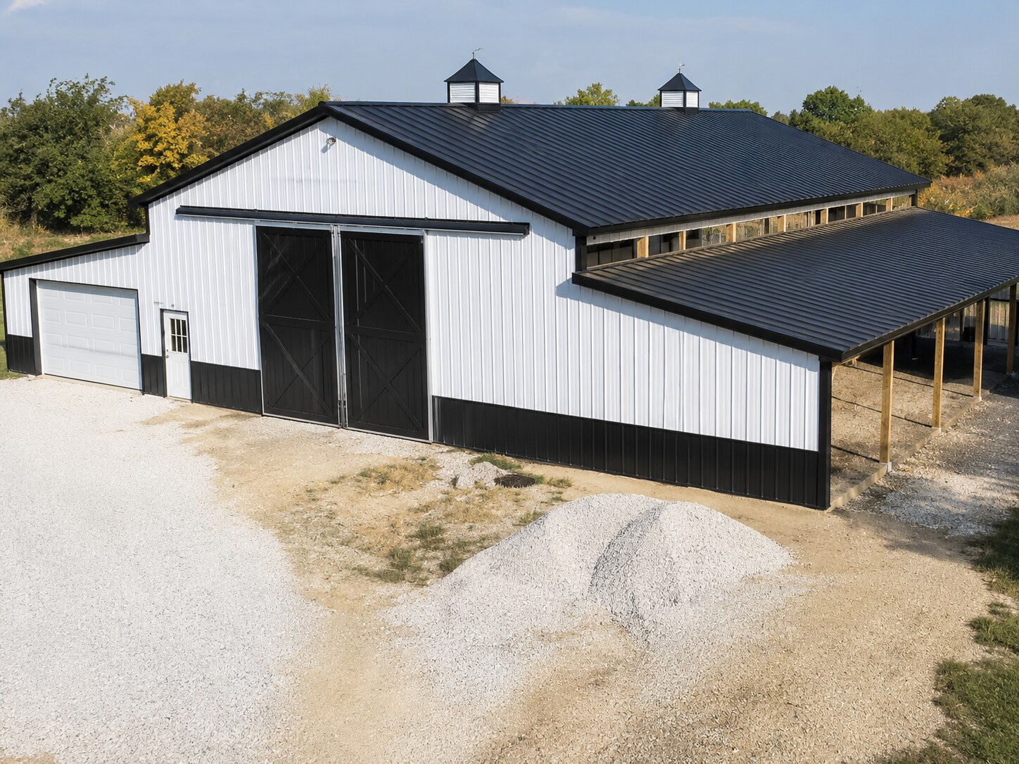 White and black post-frame barn with attached open lean-to on a gravel drive